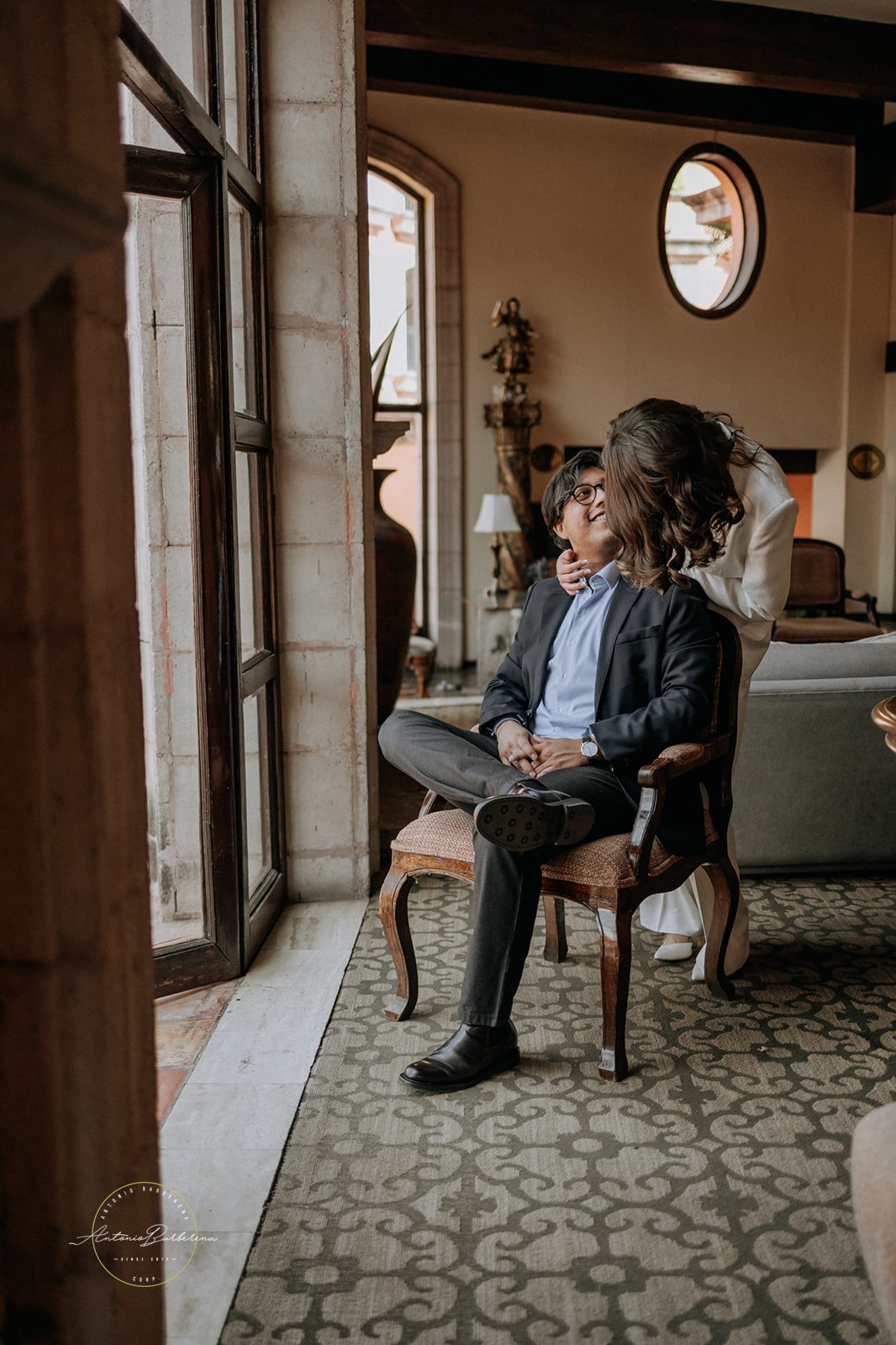 Juan Manuel sentado mientras Karla está detrás en habitación elegante